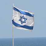 a rectangular flag, white background, blue bands close to the top and bottom, with a blue star of david in the middle. the flag is on a flagpole, with blue sky and sea in the background.