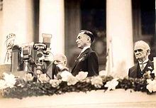 a sepia photo of a dignified debonair man on a decorated balcony who with the many microphones in front of him is about to make an address. large columns of a building are in the background and a wizened official stands at a distance behind.