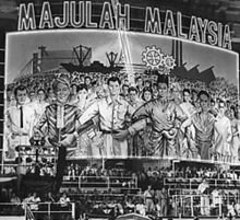 participants at a public event are dwarfed by a huge poster image displayed in the background. the poster, which is more wide than tall, depicts a crowd of confident-looking people, linking arms and looking out of the picture at the observer, with factory chimneys, a ship and other stylised industrial buildings in the background. above the image, in large letters, the words "majulah malaysia".