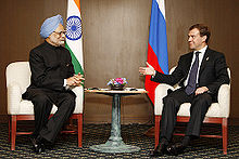 two seated men conversing. the first is dressed in indian clothing and turban and sits before an indian flag; the second is in a western business suit and sits before a russian flag.