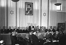 a single man, adorned on both sides by a dozen sitting men, reads a document to a small audience assembled before him. behind him are two elongated flags bearing the star of david and portrait of a bearded man in his forties.