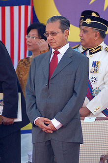 an older man with neat, slightly greying hair, dressed in a grey business suit, white shirt and red tie, his hands clasped together in front of him, wearing a pensive facial expression; behind him two men in uniform, and a well-dressed lady; the wall in the background is decorated with flags showing red and white stripes, as well as a yellow moon and star on a purple background.