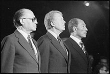 three middle-aged, suited men standing in the spotlight, seen from the side. the men are looking away from the camera, with serious expressions. in the background are several other people, out of focus.