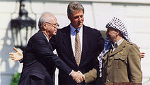 a smiling arafat, donning traditional arab headdress, shakes the hand of a stolid rabin as clinton stands tall behind and center with open arms toward the two leaders.