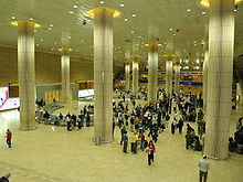 a large hall, with tall round columns; dozens of people standing or walking, around an entrance in the left wall. the area around the entrance is partitioned for the most part from the rest of the hall.