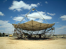 a horizontal parabolic dish, with a triangular structure on its top. around it is a flat sandy area, with desert in the background. it's a sunny day, with a few white clouds in the blue skies.